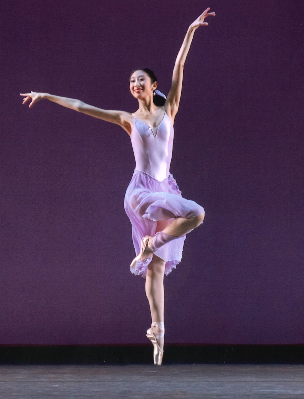 Lara Tong, in a purple dance dress, jumps with her right leg in passu00e9 and her left arm high in front of a purple stage backdrop.