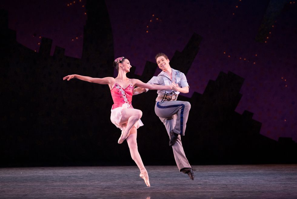 A ballerina in a pink sparkly dance dress and a male dancer in gray pants and a silver button-down shirt, hold hands and jump in passu00e9.