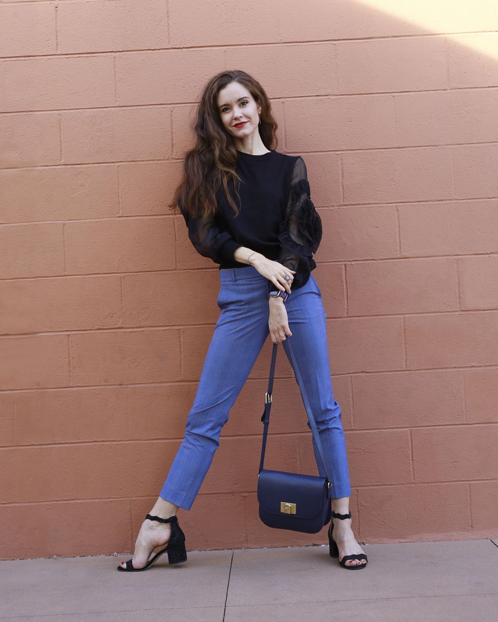 A woman with long brown hair wears a black shirt, blue plaid pants and strappy black heels and leans against a pink cinder-block wall.