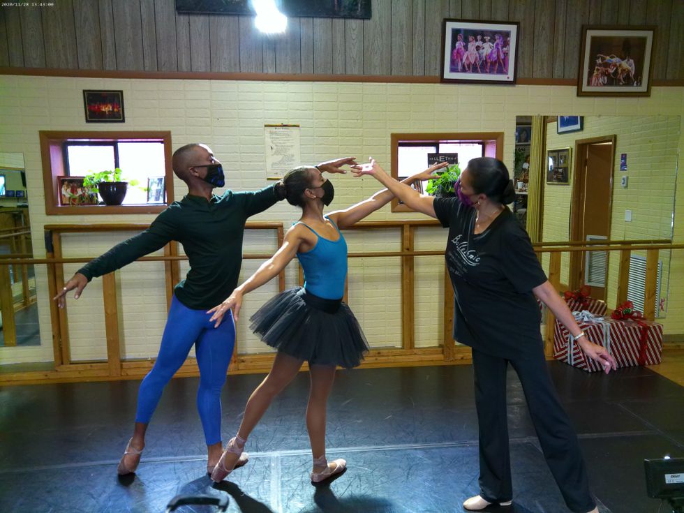 Two dancers in blue and black practice clothes and face masks, the woman in pointe shoes, pose together in a first arabesque tendu. Abarca Mitchell steps out of a mirrored pose as she adjusts the fingertips of the male dancer.