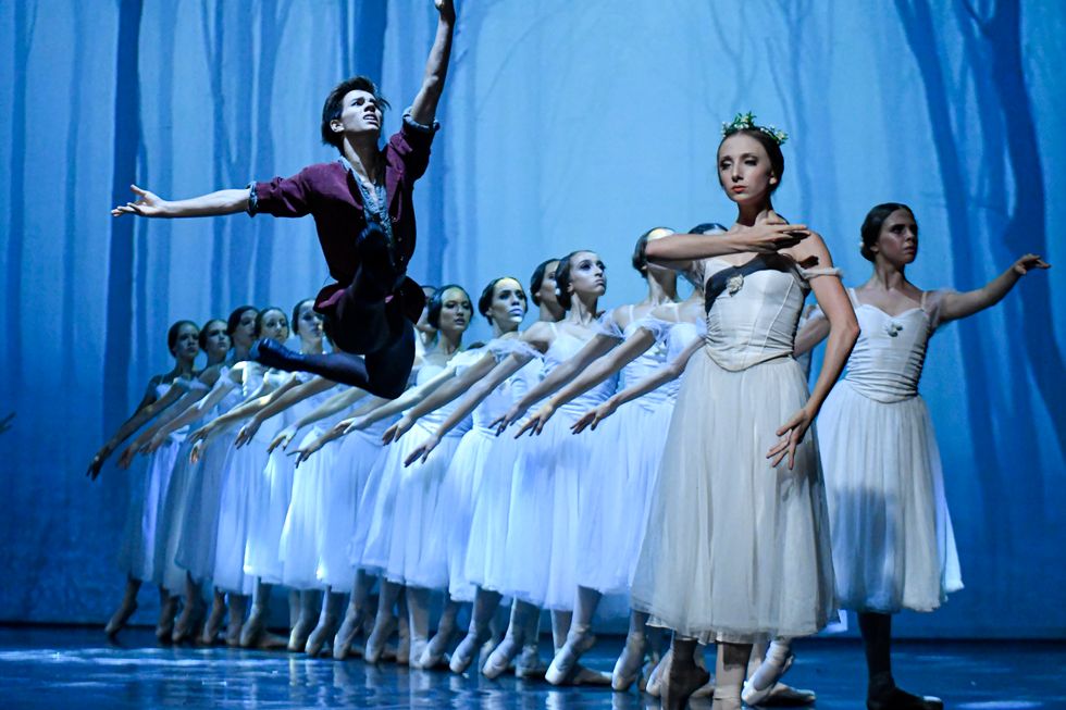 Francisco Gomes, wearing a purple peasant shirt and blue tights, leaps toward Margarida Gonçalves, who stands in tendu derriere with her right arm draped across her chest. Behind her is a long diagonal line of ballerinas in white Romantic tutus.