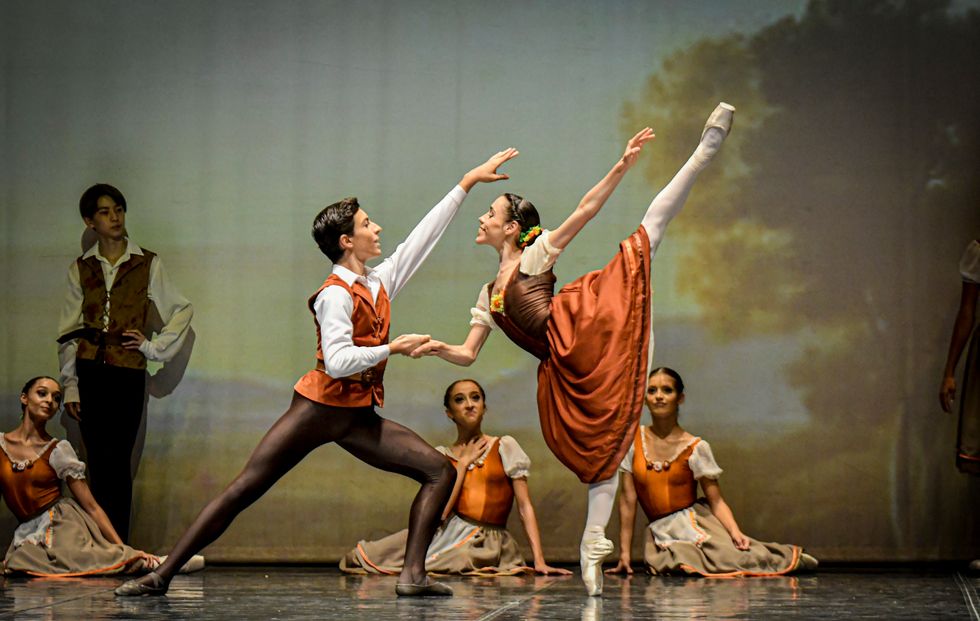 Giulio Diligente, in brown tights and an orange vest, lunges and holds the hand of Laura Viola, who, wearing an orange and brown peasant dress, does penché arabesque.