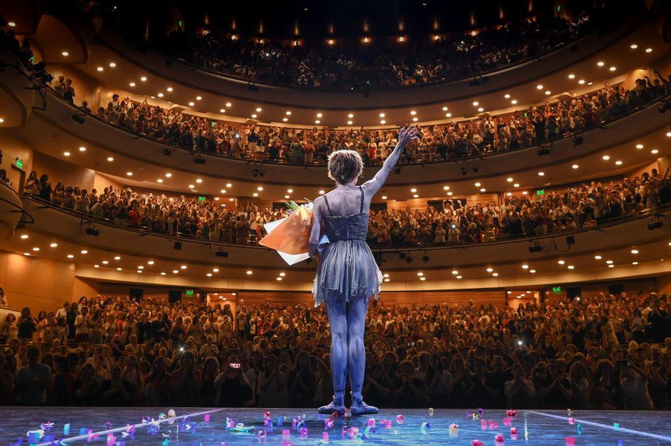 In this photo shot from behind, Maru00eda Riccetto wears the ragged dress costume and short wig from the ballet Manon and stands center stage, holding a large bouquet of flowers and waving to the audience. The packed crowd is on its feet, cheering her final bow.