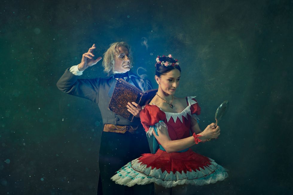 Marianela Nuu00f1ez, as Swanilda, looks at her reflection in a handheld mirror, She wears a red tutu. Behind her, Gary Avis, as Dr. Coppelius, reads a book. He wears a gray jacket and white-haired wig.