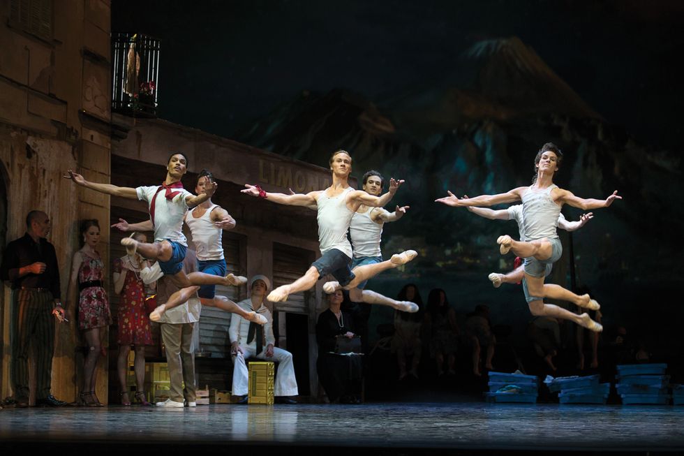 Six male dancers dressed in denim shorts and white tank tops leap in a grand jetu00e9 en avant onstage