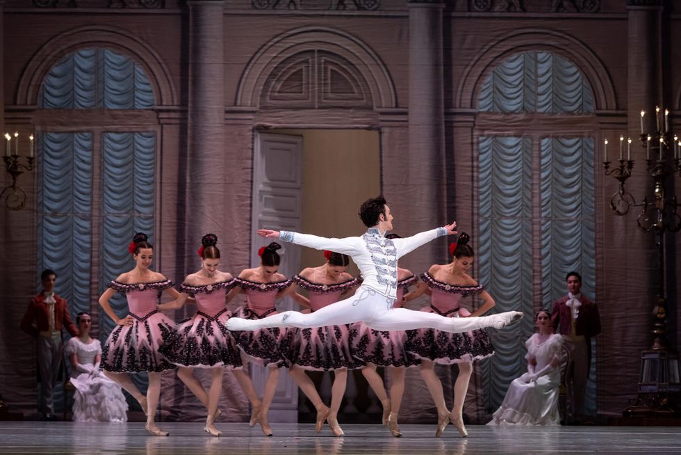 Barkidjija performs in Paquita. He is centered in the photo, on stage, in a graceful leap facing to the side. He wears white ballet boots, white tights, and a white soldier's jacket with silver embellishments. Behind him is a row of ballerinas, arm in arm, in pink tutus. Set behind them slightly are dancers dressed as courtesans, sitting and standing along the edge of the stage.
