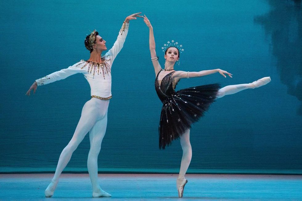 Barkidjija performs in "La Gioconda" with Maria Khoreva. Barkidjija stands to the left, with one leg in tendu derriu00e9re, and his arms in an elongated fourth position. He faces Khoreva, wearing white tights, white ballet shoes, a white long sleeved tunic embellished with gold, and a gold crown. Khoreva stands in arabesque on pointe, facing the audience, her higher arm extended towards Barkidjija. She wears pink tights, pink pointe shoes, a black tutu embellished with silver, and a silver tiara.
