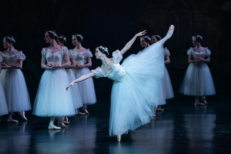 Bianca Scudamore, a pale white woman dressed in a white Romantic tutu and pointe shoes, balances in an extended first arabesque line, torso diving towards the floor. Behind her, a corps of identically dressed women pose in neat lines in B-plus, hands crossed at the wrist.