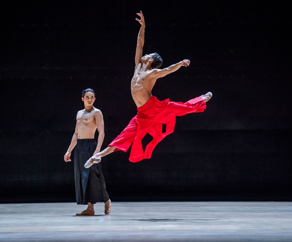 A lean, bare-chested man leaps into the air. His front leg is at a 45 degree angle to the ground, the back one in a perpendicular attitude. His head tips to the ceiling as his right arm reaches gracefully upward, the left outflung to his side. The bright red trousers he wears flare with the motion. Upstage, another man costumed identically but for the black color of his trousers looks on.