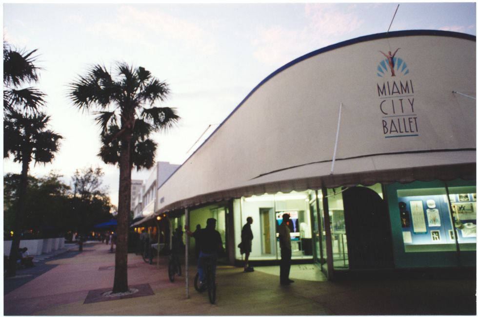 A palm-tree-lined street scene at dusk shows a white one-story storefront with Miami City Ballet written on the facade and people milling about outside.
