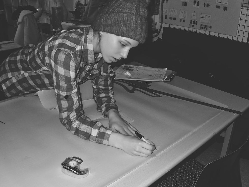 IN this black-and-white photo, a young woman in a plaid shirt and knit hat leans over a table to sketch a design with a pen.