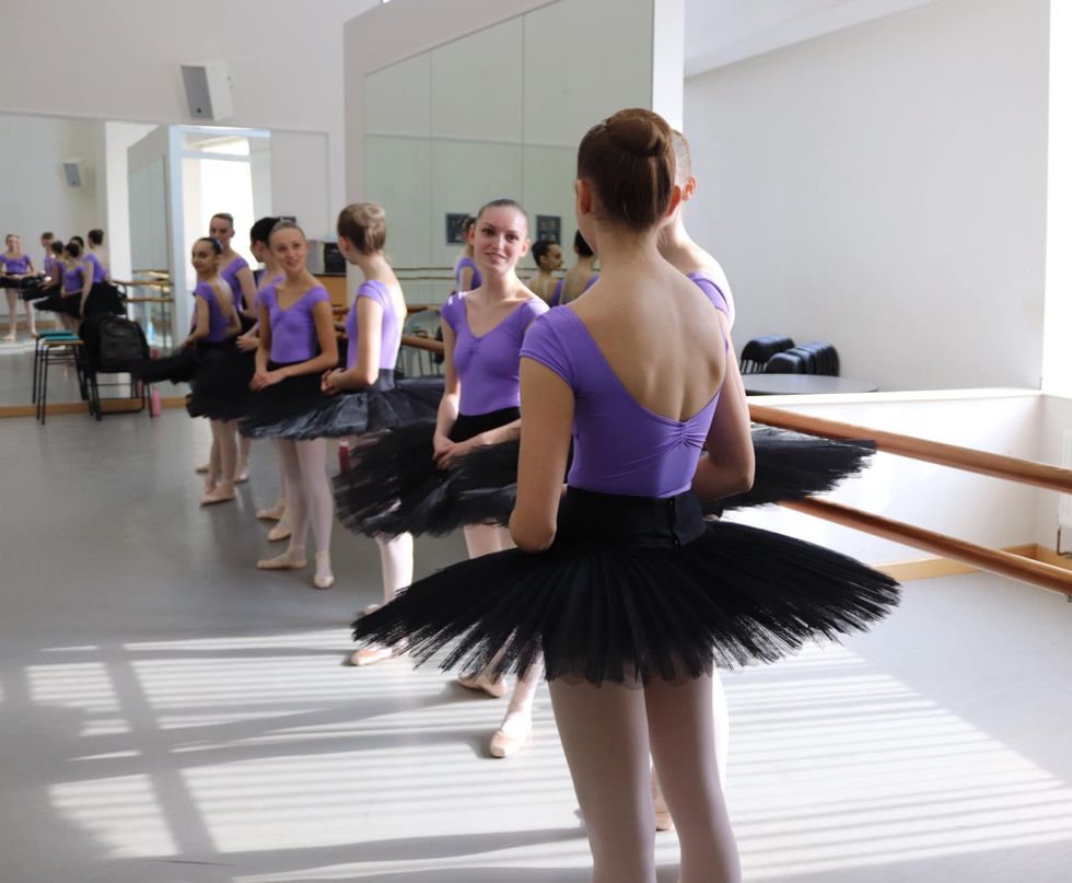 Twelve female ballet students stand casually in a sunny dance studio, wearing purple caps-leeved leotards, pink tights, pointe shoes and black practice tutus.