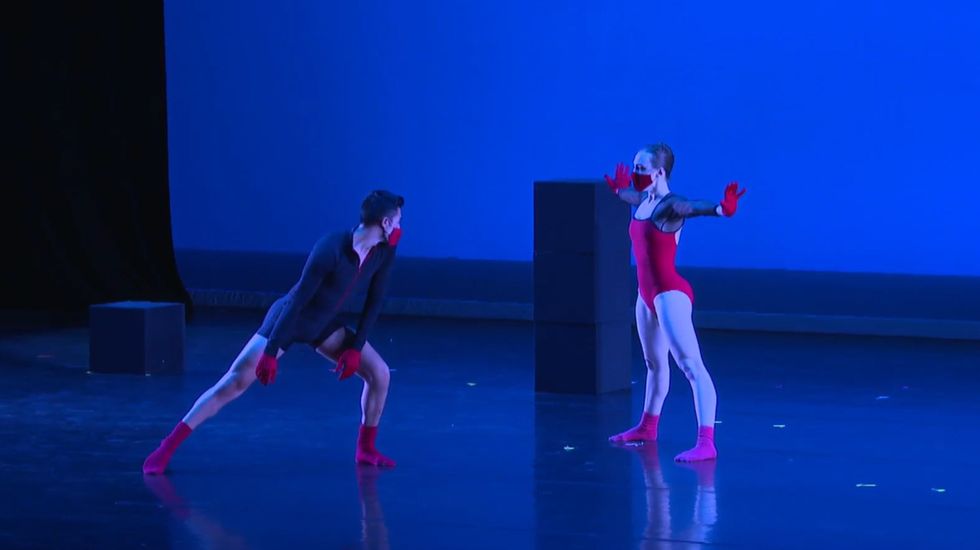 A male and female dancer face each other onstage wearing red socks, gloves and a masks. The woman, in a red leotard, stands with her feet apart and her arms wide and hands flexed while the man, in a black leotard, lunges toward her on his left foot.