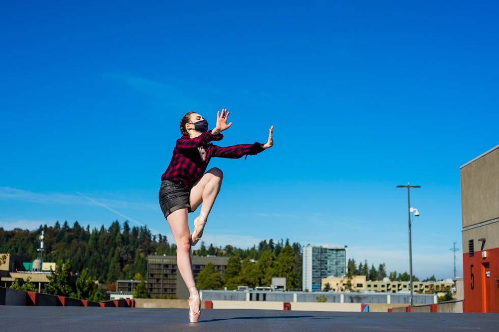 Wearing a red and lack plaid shirt, jean shorts and pointe shoes, Kyra Laubacher stands on a rooftop and does a parallel passu00e9 with her left, pushing her arms forward with flexed hands.