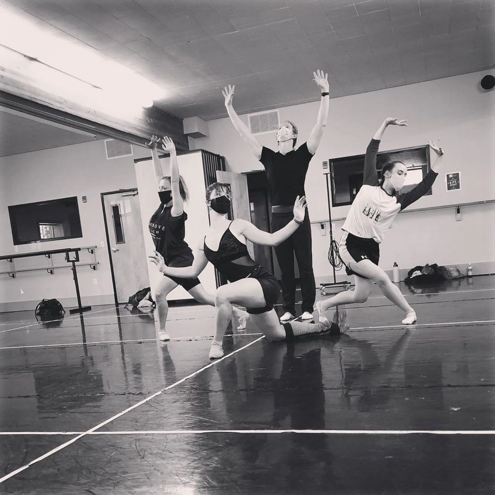 A male dancer stands in the center of a studio with his arms held up, with three female dancers crouched around him.