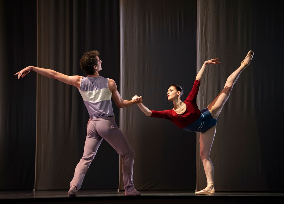 A male dancer in a purple costume faces a ballerina in a red and blue leotard and holds her right hand as she takes arabesque allongu00e9 in pliu00e9.