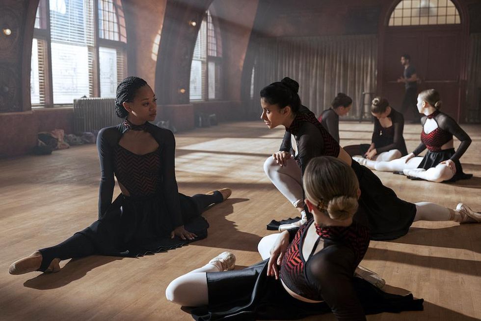 A still from "Tiny Pretty Things." Dancers stretch on the floor of the studio. Our focus is on Kylie Jefferson and Daniela Norman, who seem to be engaged in conversation while stretching. Both girls look serious, and face each other. Kylie Jefferson is in a split, and Daniela Norman is in a lunge. Both wear long-sleeved dresses, tights, pointe shoes, and their hair in buns.