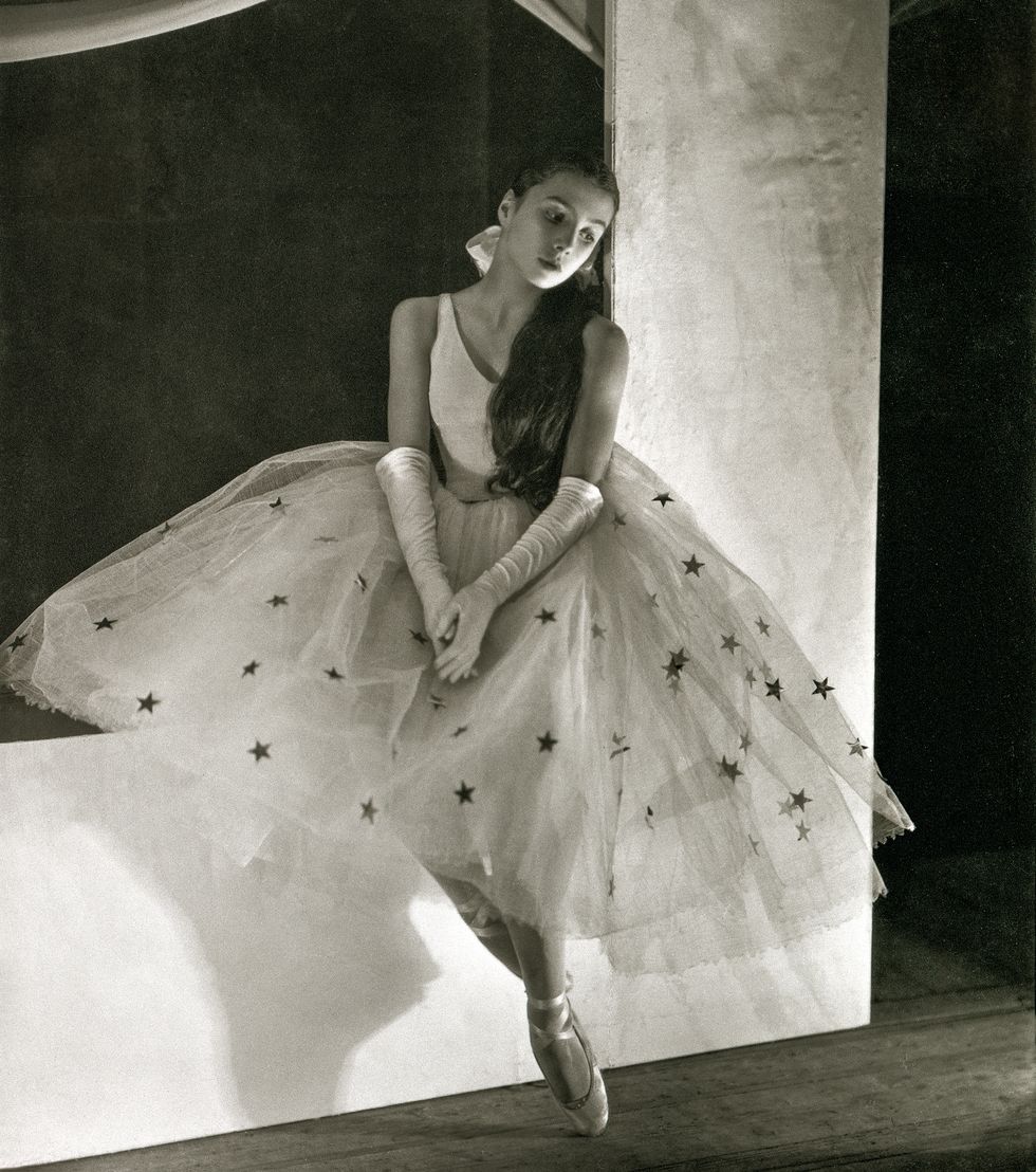 A black and white photo of Toumanova in a large tulle tutu covered in silver stars, white bodice and white gloves sitting on a large white set piece with her hands in her lap.