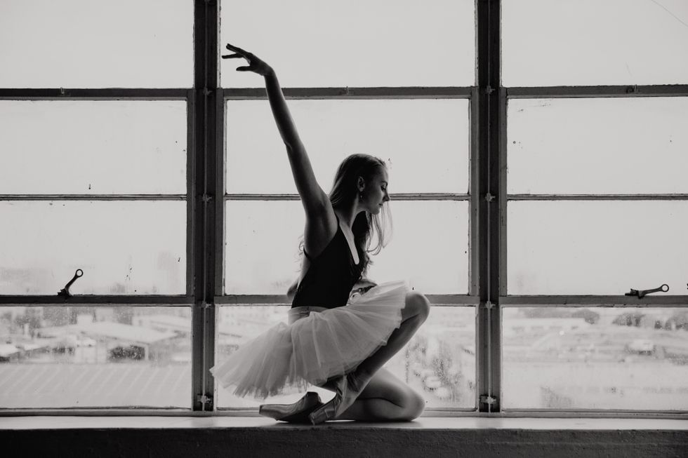 Black and white photograph of Lebowski in a black leotard and white tutu kneeling against a large window in profile. She has one arm lifted.