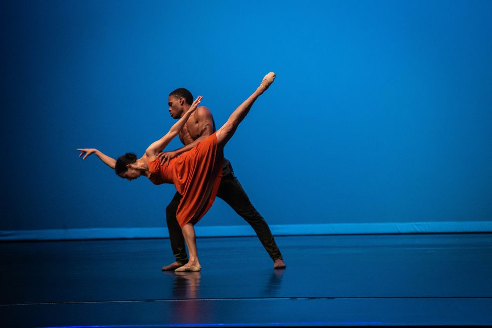 Hill is in a high arabesque penchu00e9 with her face parallel to the floor. She wears an orange dress and ballet slippers, and is held under her armpits by Vessell, who is standing in a lunge and wearing black pants and no shirt.