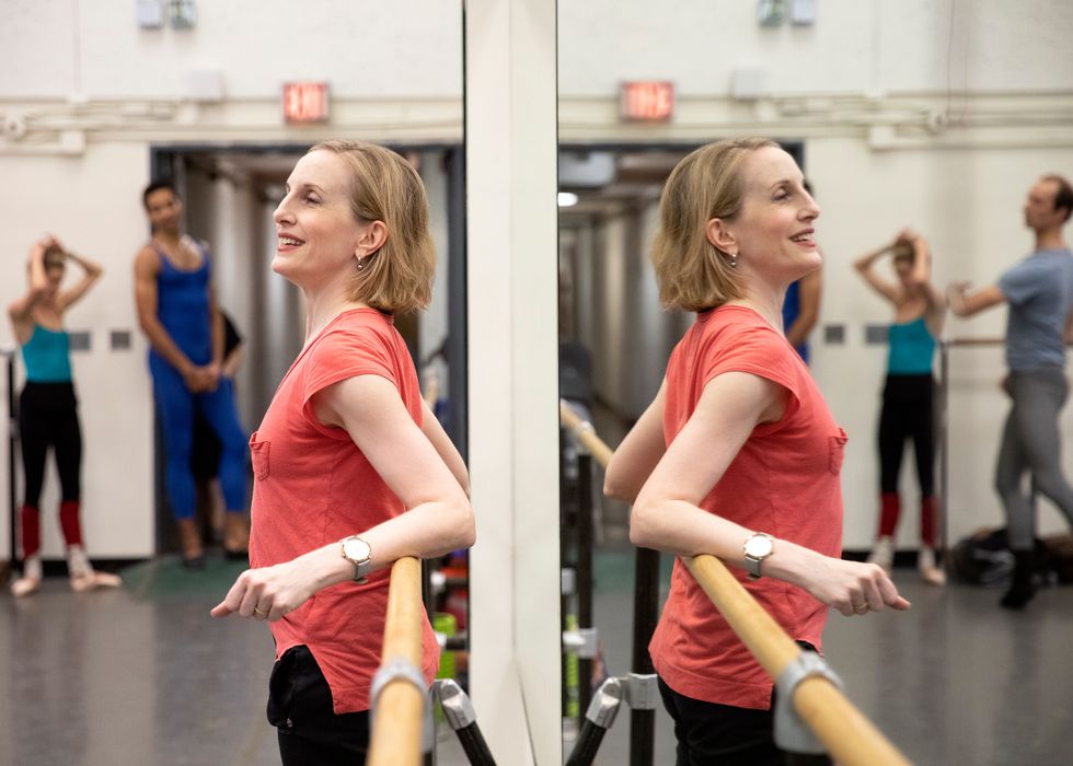 Wendy Whelan, in an orange t shirt, leans against the barre and mirror in a ballet studio.
