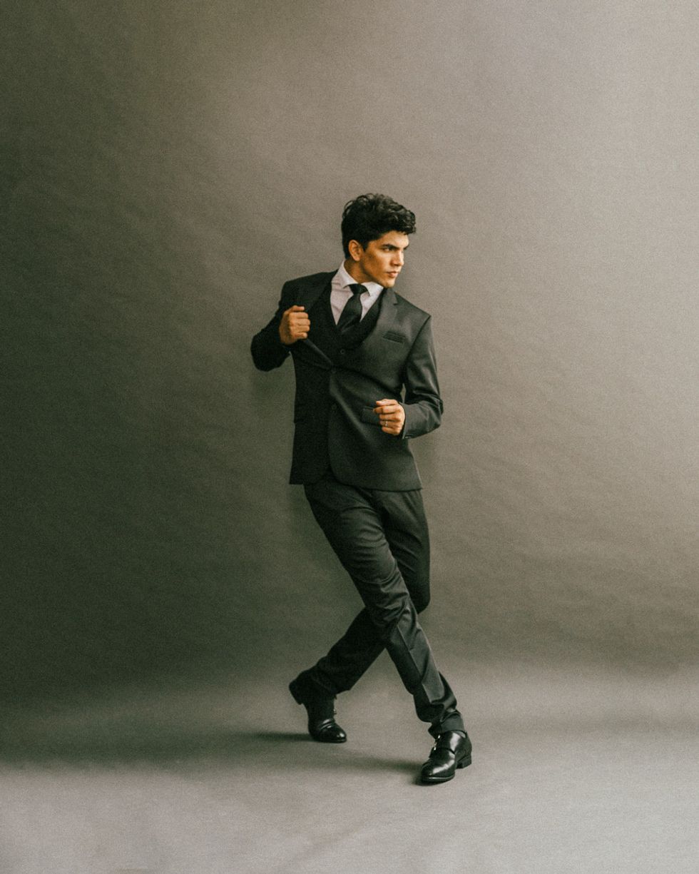 A male dancer in a gray suit and tie tugs at his right lapel and crosses his right foot in front while popping his left foot up.