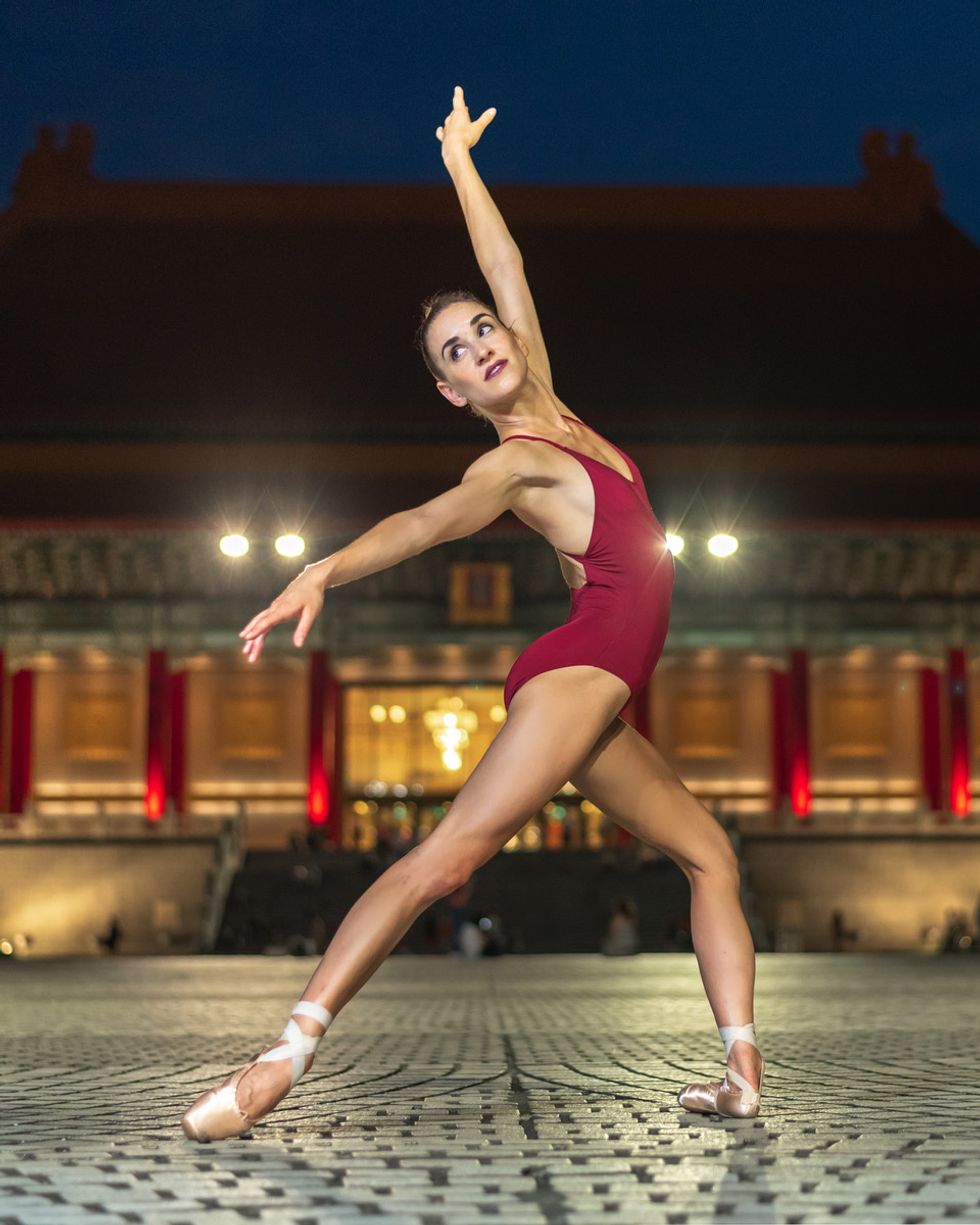 A female ballerina in a maroon leotard stands on an outdoor stage in tendu deriu00e9rre, looking back over her right shoulder.