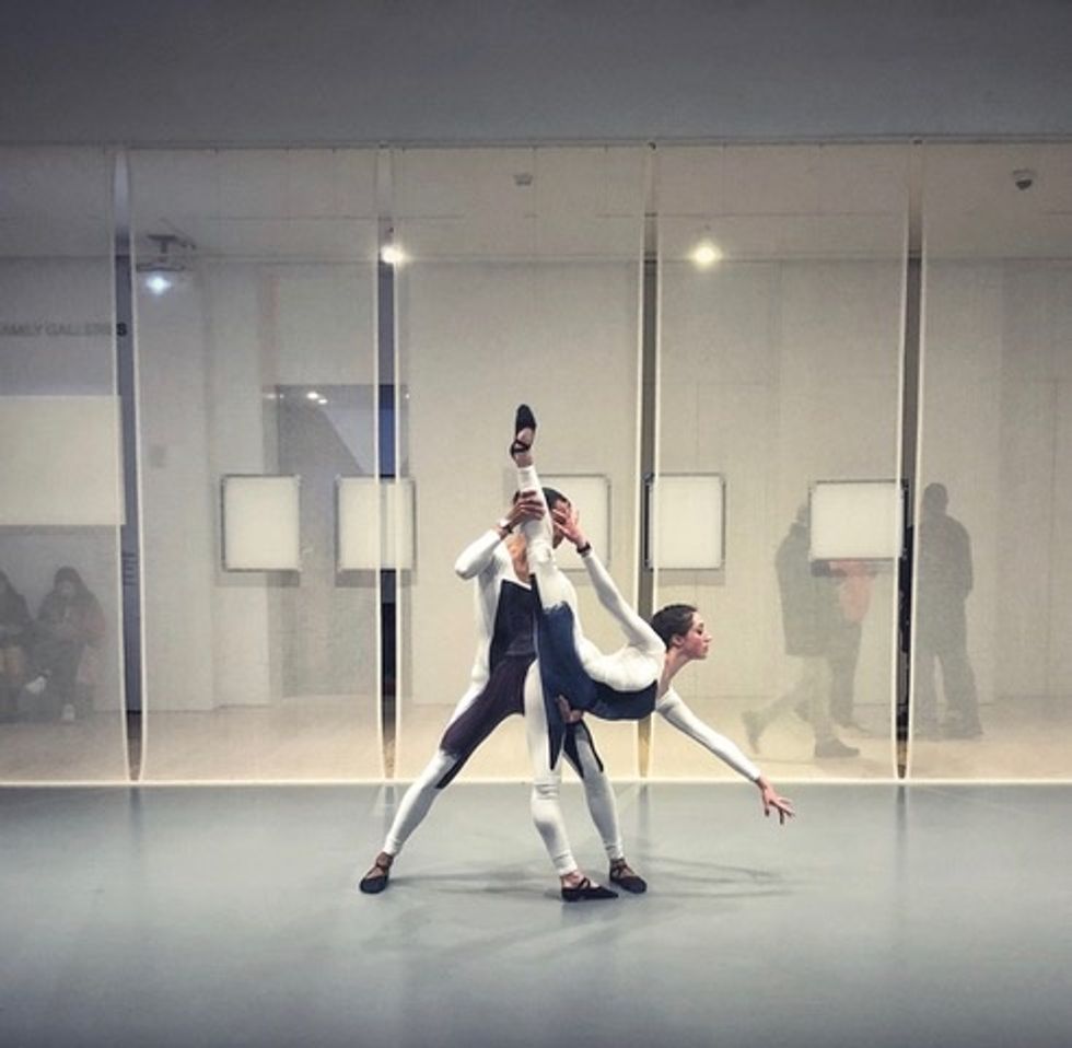 Evelyn Kocak, wearing a white and blue unitard and ballet slippers, takes penchu00e9 while her partner (wearing the same costume) holds her back leg