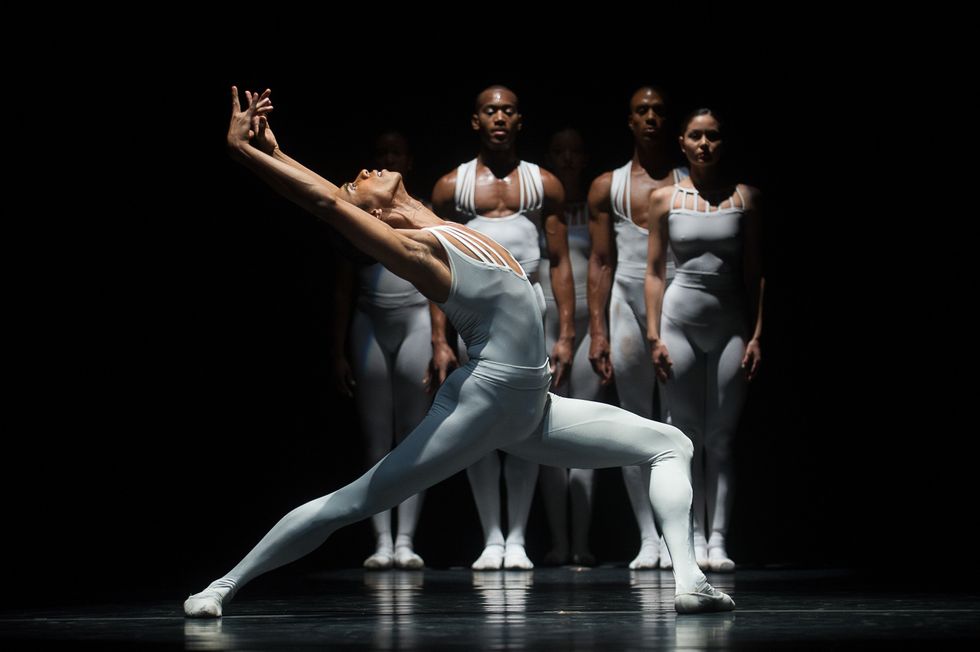 Wearing a white unitard, Francis Lawrance lunges on his left leg in profile and bends backward with his arms in high fifth position. He stands in front of several men in women in similar costumes on a darkened stage.