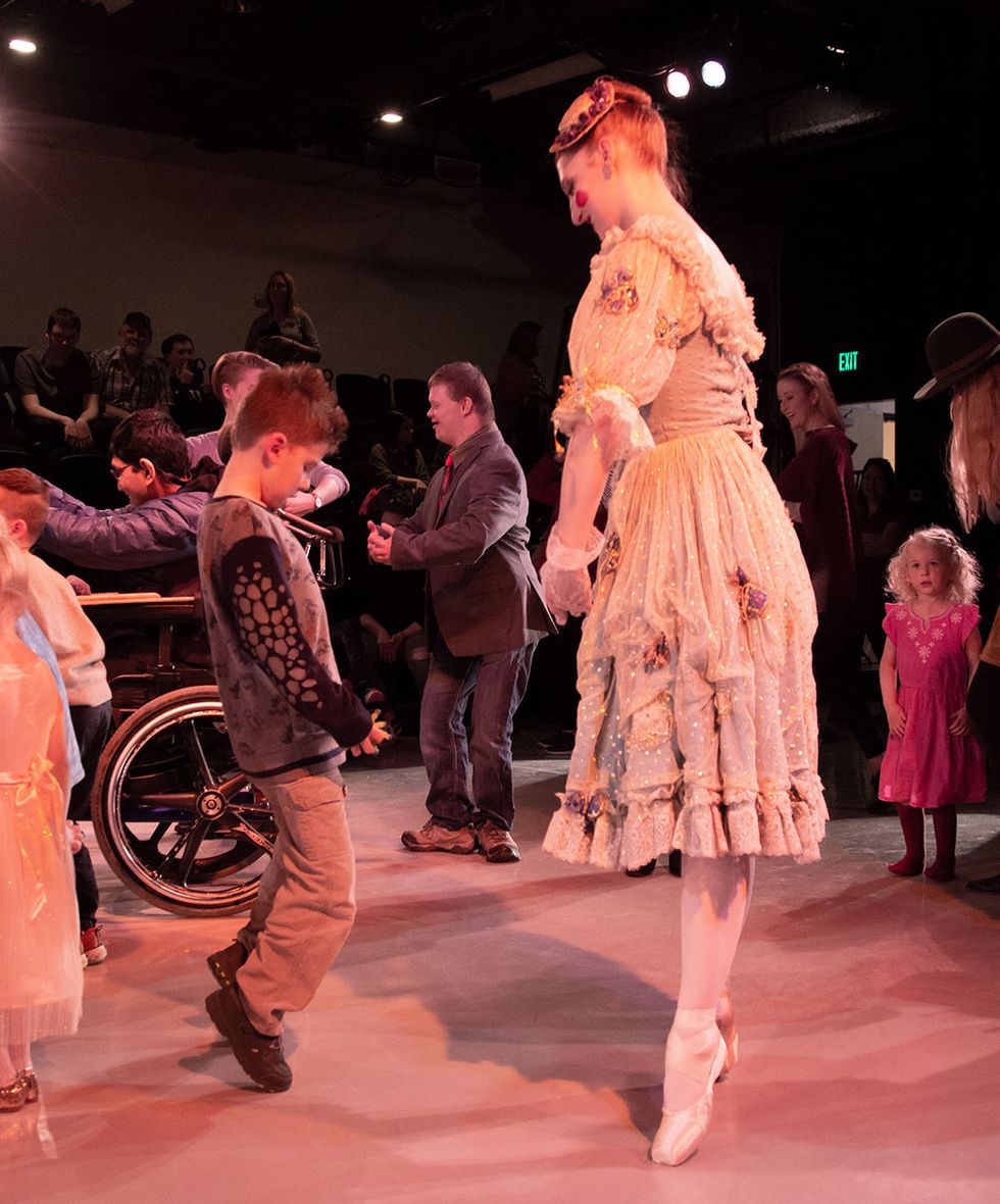 A ballerina demonstrates standing on relevu00e9 to a young boy. They're surrounding by other children and adults practicing dance moves.