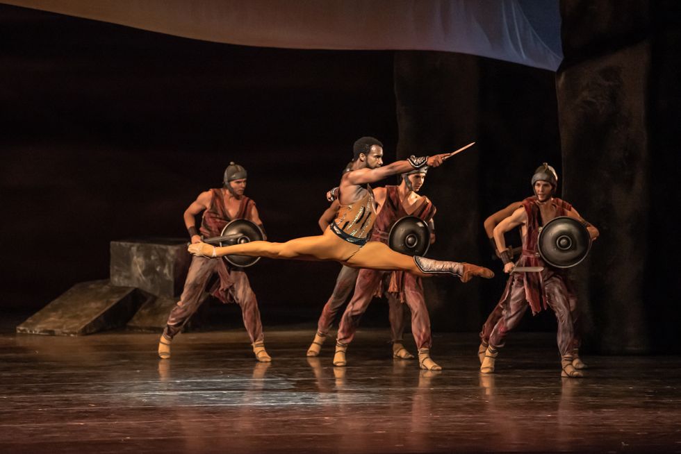Wearing orange tights and vest and gold dance boots, Osiel Gouneo leaps to the left and extends his right arm forward, holding a dagger.