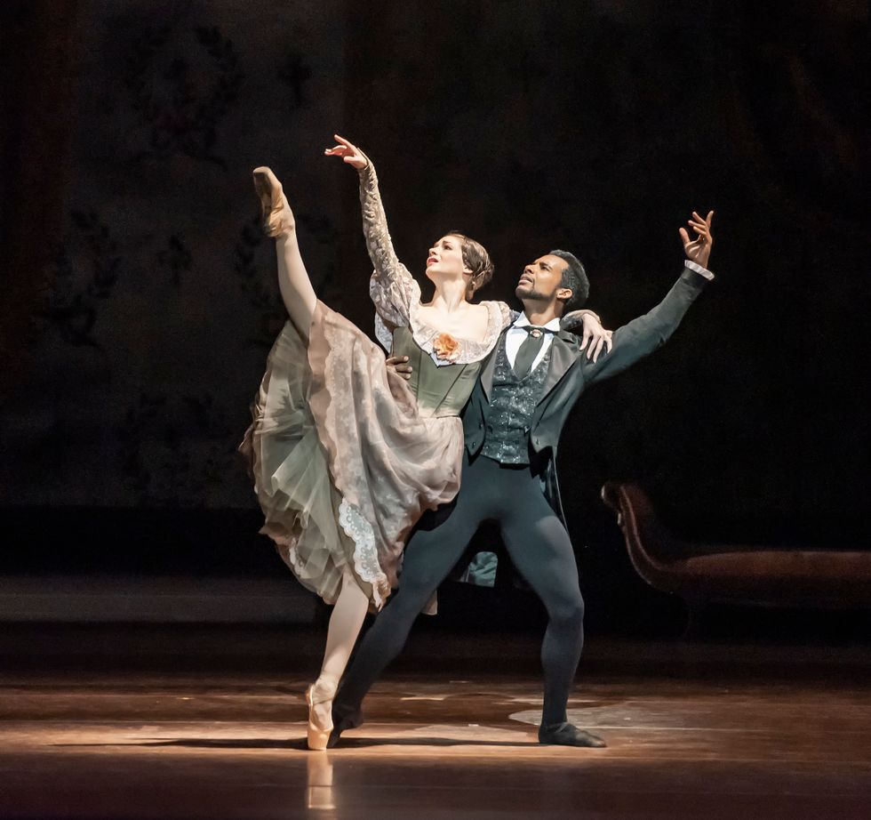 Osiel Gouneo wears a black jacket, vest and tights and lunges back on his left leg while holding ballerina Laurretta Summerscales by the waist. Summerscales, in a long-sleeved green and pink 19th century dress, leans against him and does a developpu00e9 with her right leg.