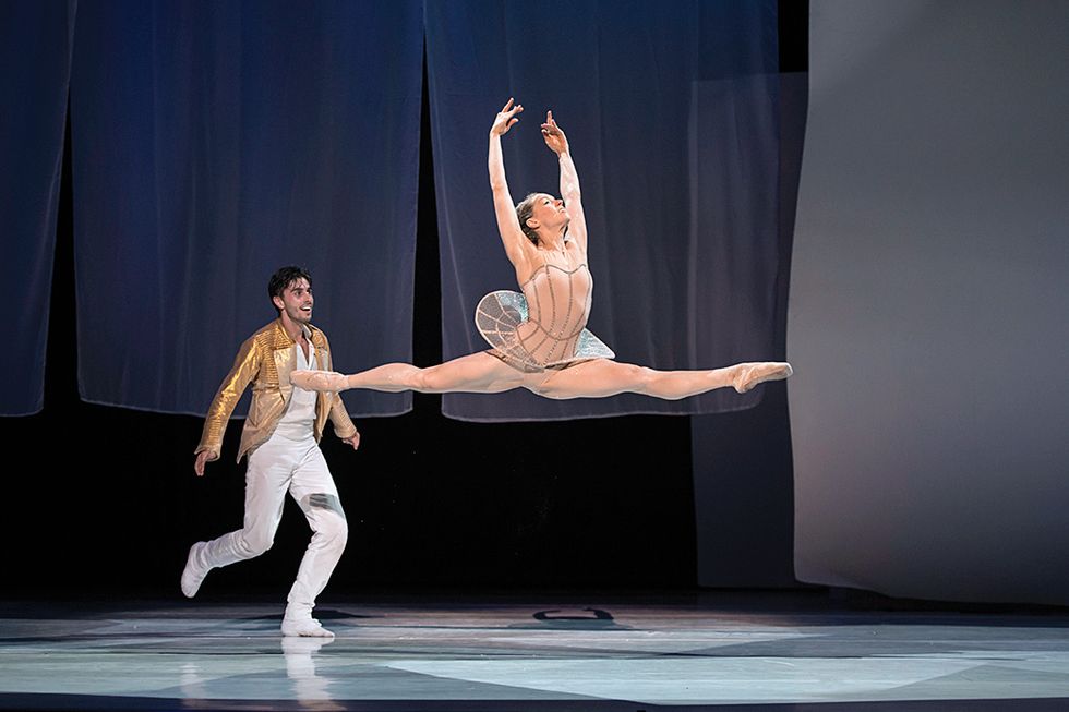 Ball onstage in a sheer bodice and skirt leaping with her arms in fifth position, while a man in white pants and a gold jacket chases behind her.