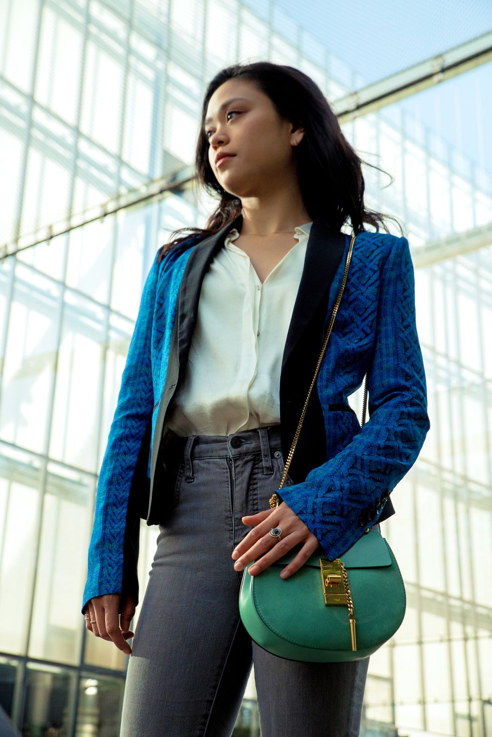 Angelica Generosa looks off to her right in front of a glass-windowed building. She wears a blue blazer, white blouse, gray jeans and carries a small green handbag.