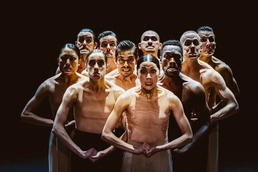 A group of 10 dancers stand in a pyramid shape with their hands clasped tightly and their lower jaws thrust out. They wear flesh tops and taupe and black pants.