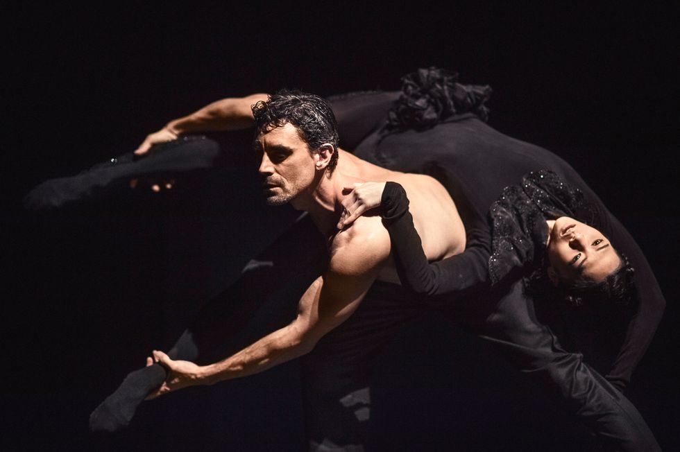 A shirtless male dancer shifts a woman in black tights and embellished black leotard across his back, holding on to both her ankles.