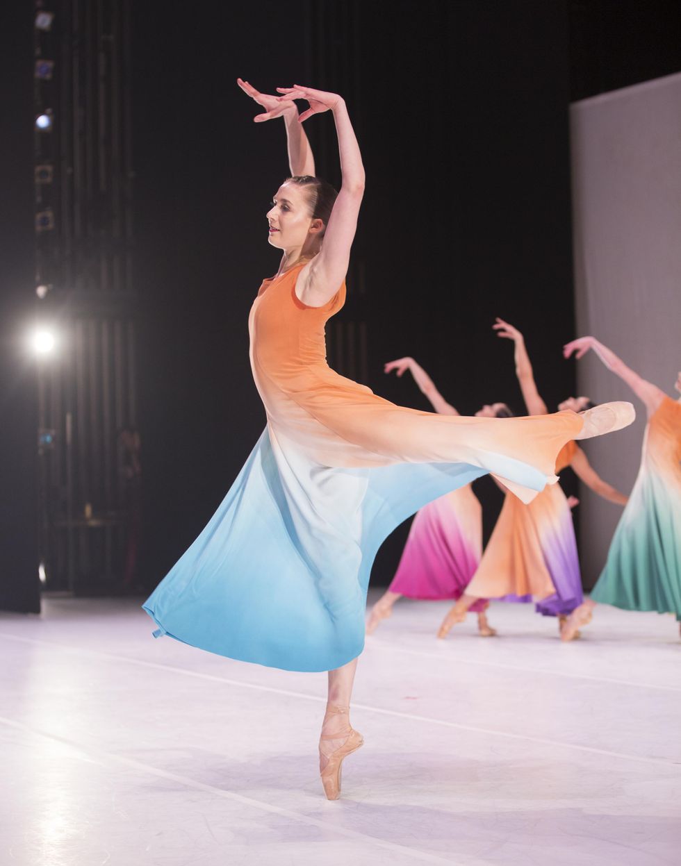 Cecilia Iliesiu in attitude derriu00e8re on pointe. She is wearing a pastel-hued dress that floats with her movement. Her arms are extended above her head.