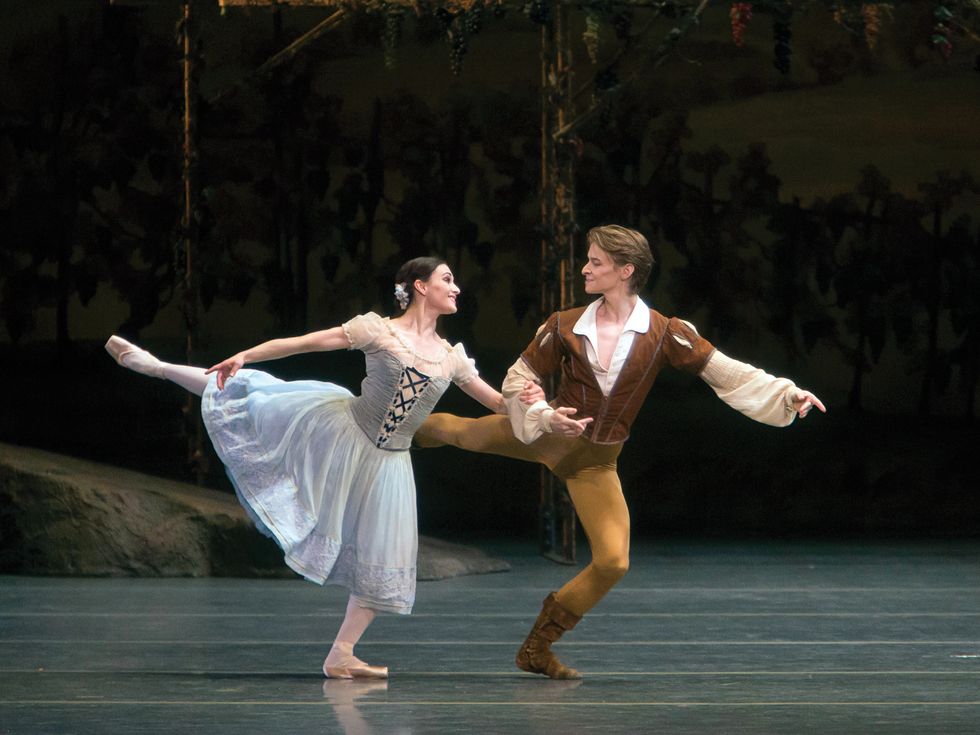 Simkin, dressed in a white blouse and brown vest and tan tights and boots, hooks arms with Lane, dressed in a blue peasant dress, as they chug along in an arabesque with their front knees bent and look at each other.