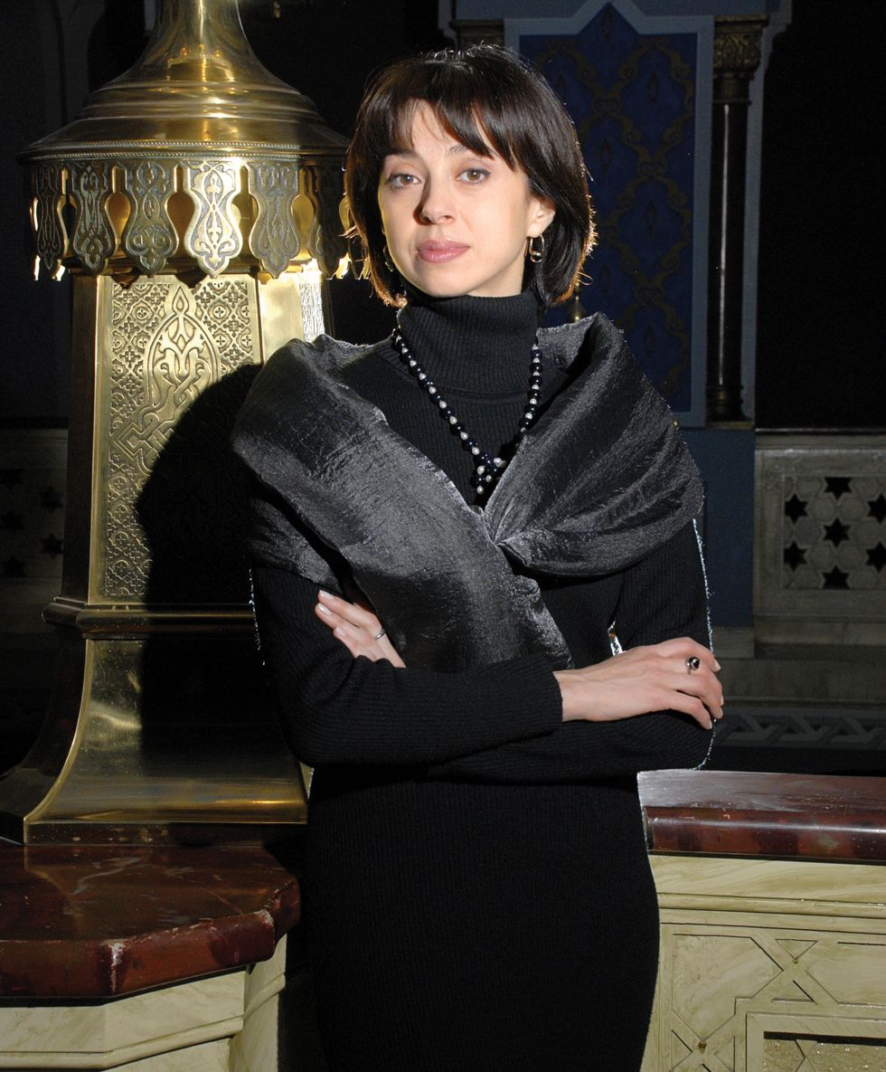Ananiashvili leaning against a regal background in black sweater and gray silk scarf with her arms crossed