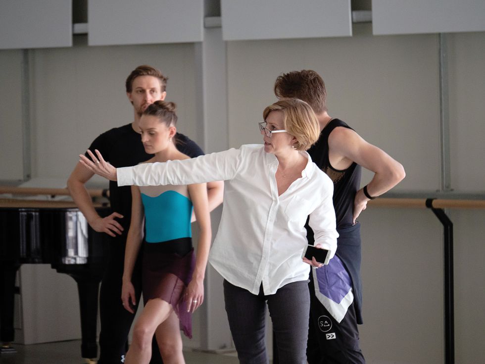 Barker, in a white blouse and jeans, directs a rehearsal in a studio with three dancers standing behind her