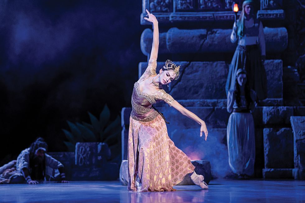 Semionova in a pink and gold outfit and crown, in a deep pliu00e9 onstage with one leg behind her and her arms framing her face.