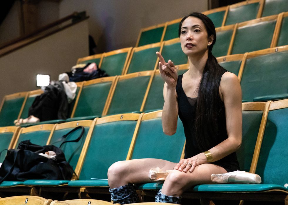 Abrera sits in a green theater seat, pointing at the stage while sewing pointe shoes in her lap.