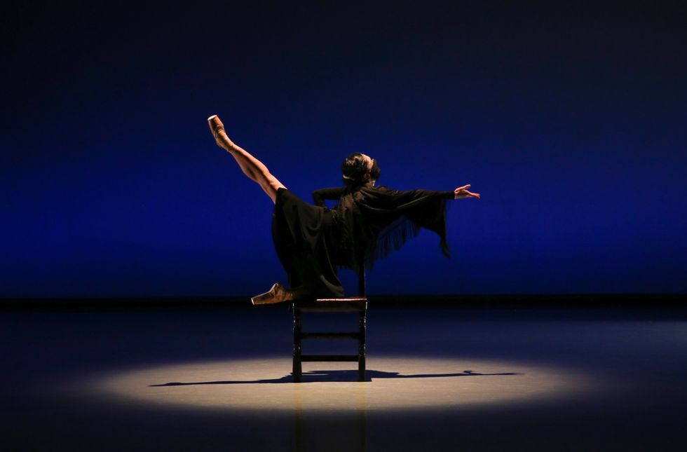 â€‹Eri Nishihara, in a black dress and face mask, balances on a chair, kneeling in arabesque while reaching forward with her right hand.