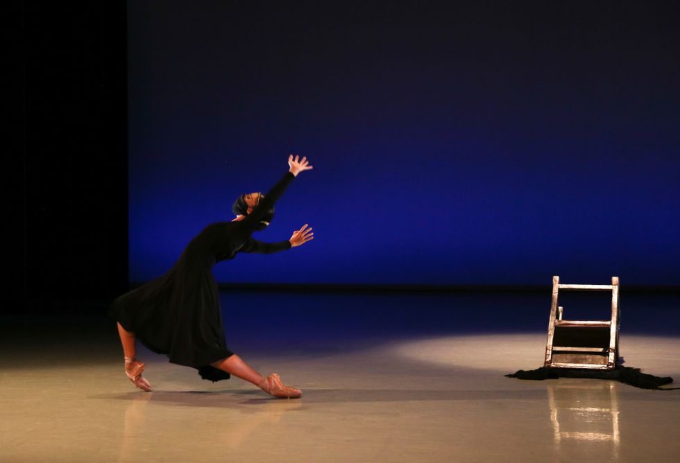 â€‹Elena Bello, in pointed shoes, a black dress and a face mask, slides on pointe across the stage. A chair is overturned to her right.