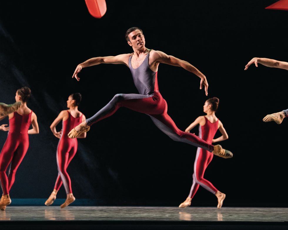 Diego Crus, in a red and purple unitard, leaps with both legs in attitude and his arms sloping down.