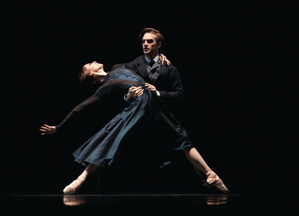 On a dark stage, Thatcher, dressed in a black suite, holds Stahl, dressed in a blue and black dress, as she stands on pointe in a wide fourth position and leans back.