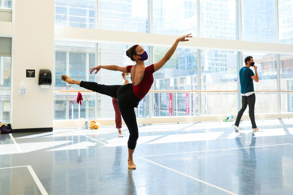 Anais Buenou2014wearing a red leotard, black tights, tan slippers and a black face masku2014performs a fourth arabesque on her right leg. She dances in a bright studio surrounded by large windows.