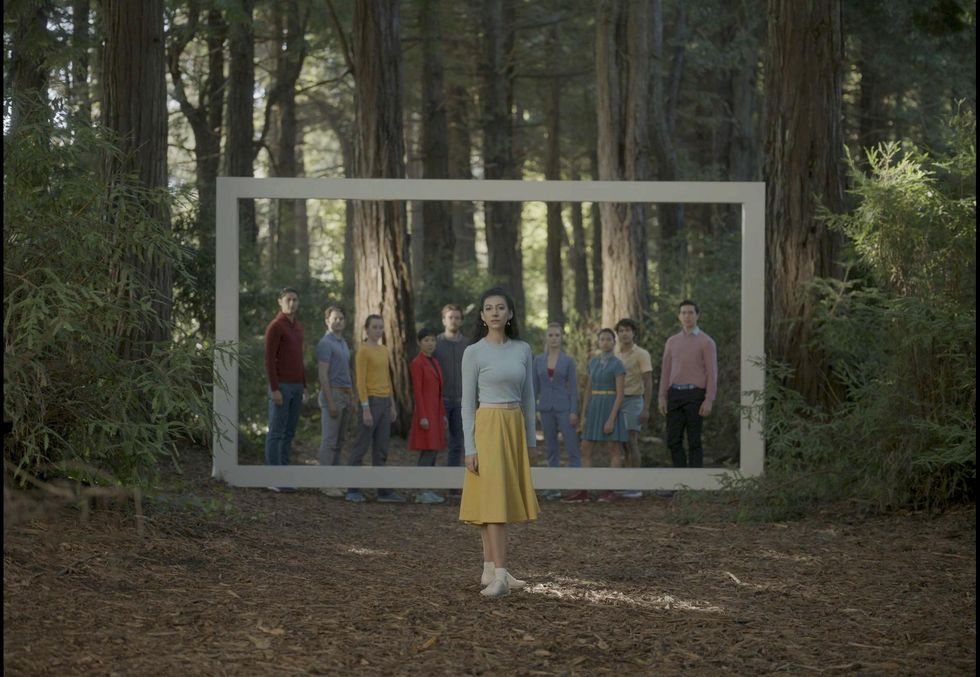 Within a forest, a group of dancers stand casually behind a large, rectangular-shaped metal frame. A woman with dark brown hair, yellow skirt and off-white long-sleeved top stands in front of them and looks straight ahead.