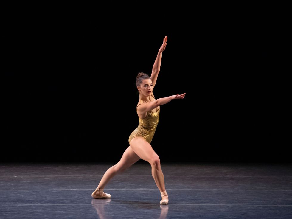 Indiana Woodward wears a gold leotard and stands in a large fourth position lunge onstage.