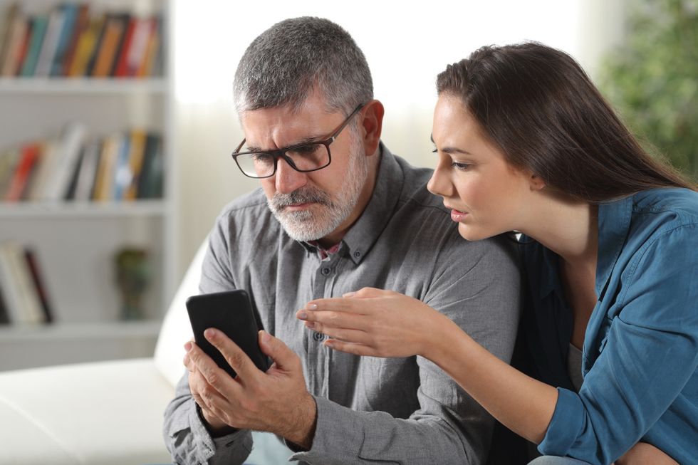A father with glasses and a gray beard holds a phone in his hand, frowning, while his daughter leans over his shoulder.